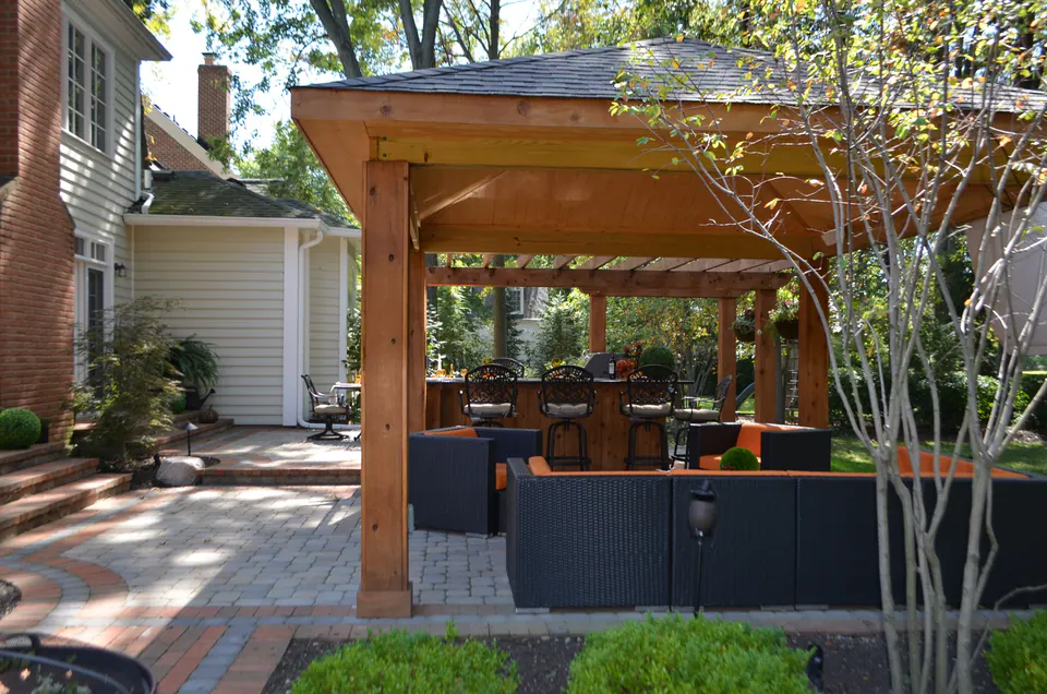Covered backyard pavilion with lounge seating, paver patio, wood posts, house connection, and planting beds.